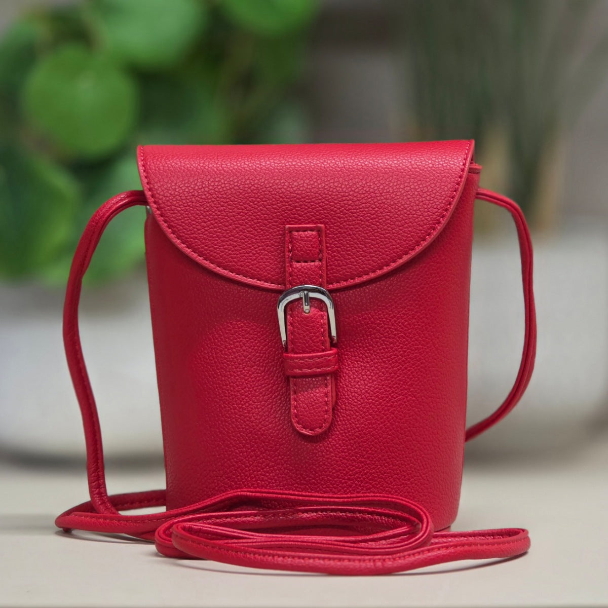 A bright red pebbled leather crossbody bag with a flap closure, silver buckle, and matching shoulder strap, shown against a softly blurred green and gray background.