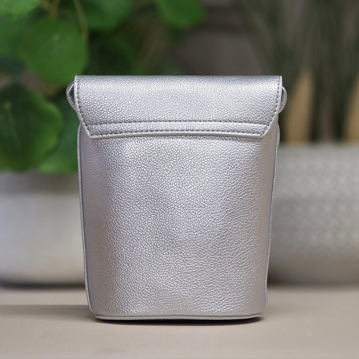 A silver textured leather pouch with a flap top, standing upright on a neutral surface with a blurred background of green leaves and a gray container.