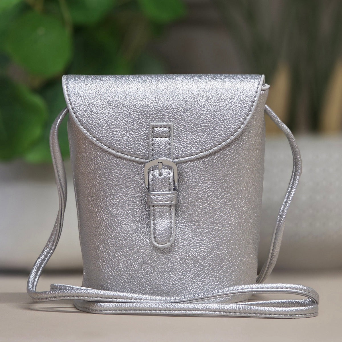 A silver textured faux-leather crossbody bag with a flap closure, front buckle strap, and thin shoulder strap, shown against a softly blurred background with green foliage.