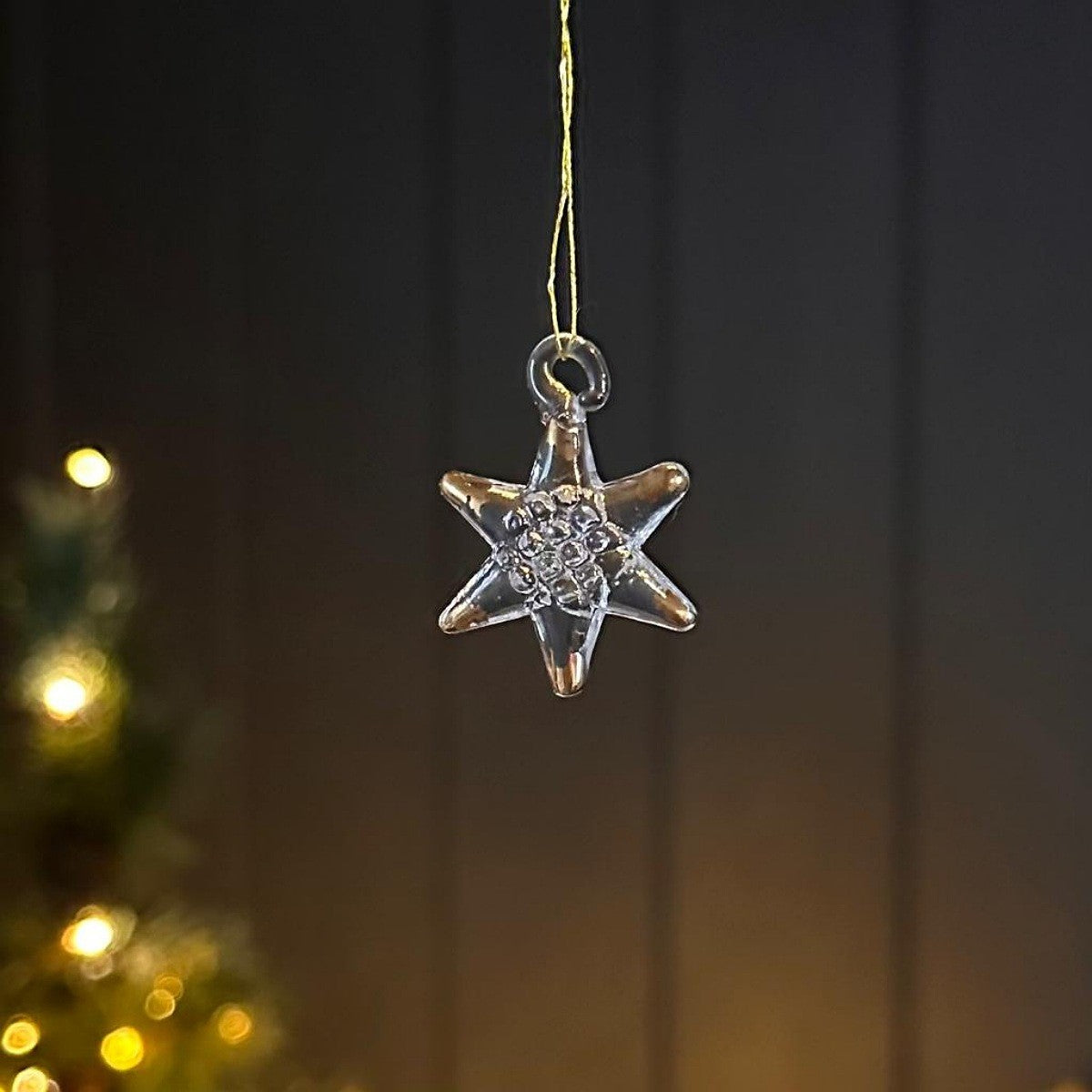 A clear glass six-pointed star Christmas ornament with gold effects hanging from a thin gold string against a dark, softly blurred background with warm tree lights.