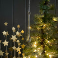 A clear glass teardrop-shaped hanging ornament suspended in front of a lit Christmas tree, with gold star decorations on the left against a dark paneled background.