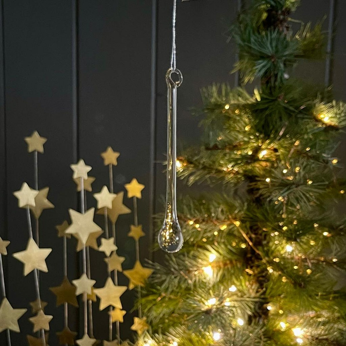 A clear glass teardrop-shaped hanging ornament suspended in front of a lit Christmas tree, with gold star decorations on the left against a dark paneled background.
