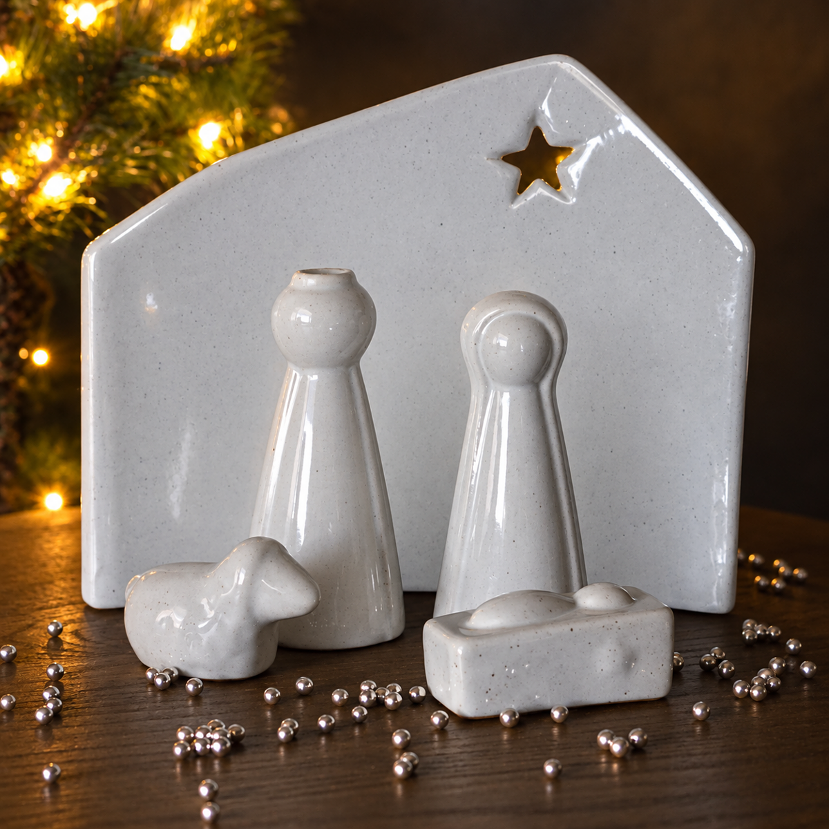 A white speckled ceramic nativity scene set on a tabletop with small metallic shiny balls scattered around. The scene includes a ceramic arched stable with open star a couple of figures a small cradle piece and a sheep.