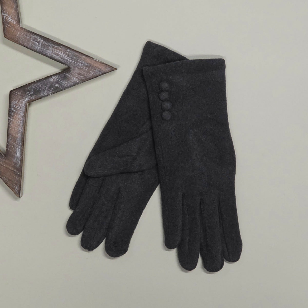 A pair of black felt gloves laid flat on a light beige background, with one glove showing four decorative buttons and a small floral detail on one fingertip. A wooden star-shaped prop is partially visible at the top left.