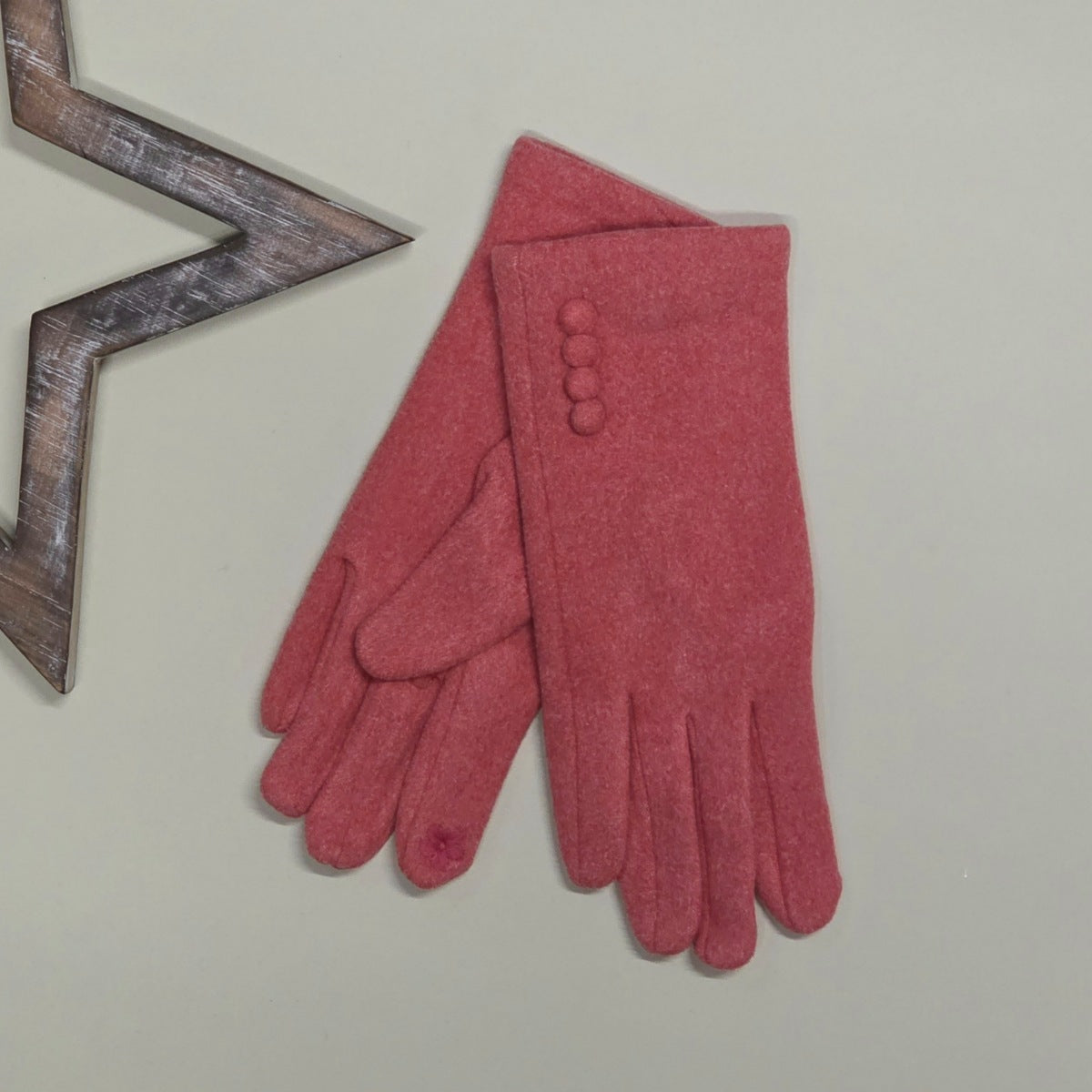 A pair of coral felt gloves laid flat on a light beige background, with one glove showing four decorative buttons and a small floral detail on one fingertip. A wooden star-shaped prop is partially visible at the top left.