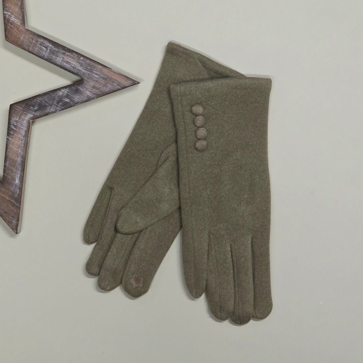 A pair of green felt gloves laid flat on a light beige background, with one glove showing four decorative buttons and a small floral detail on one fingertip. A wooden star-shaped prop is partially visible at the top left.