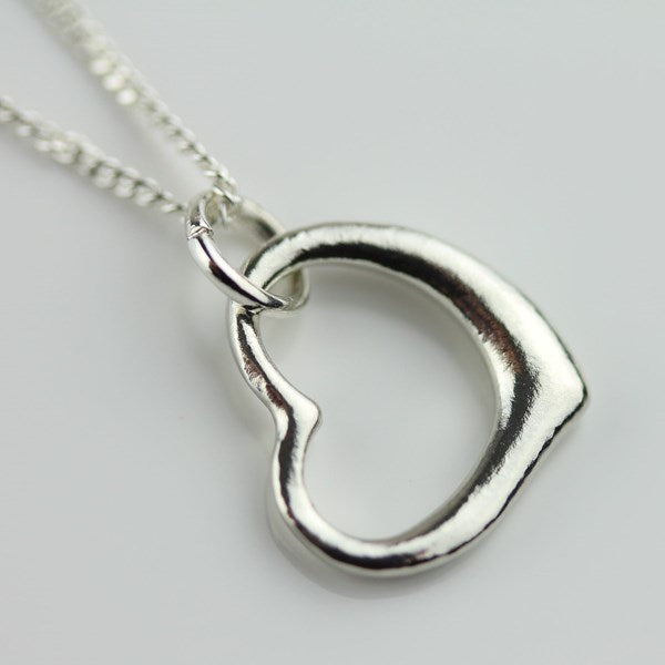 A polished silver pendant necklace featuring an open, textured heart-shaped design on a matching silver chain, photographed against a neutral light gray background.