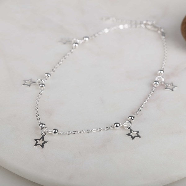 A delicate silver anklet featuring a fine chain with small silver beads and three dangling star charms, displayed on a light marble surface.
