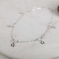 A delicate silver anklet featuring a fine chain with small silver beads and three dangling star charms, displayed on a light marble surface.