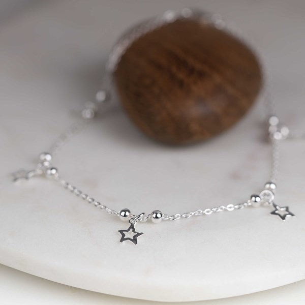 A delicate sterling silver anklet featuring three small star charms and polished bead accents, displayed on a white marble surface with a dark wooden stone in the background.