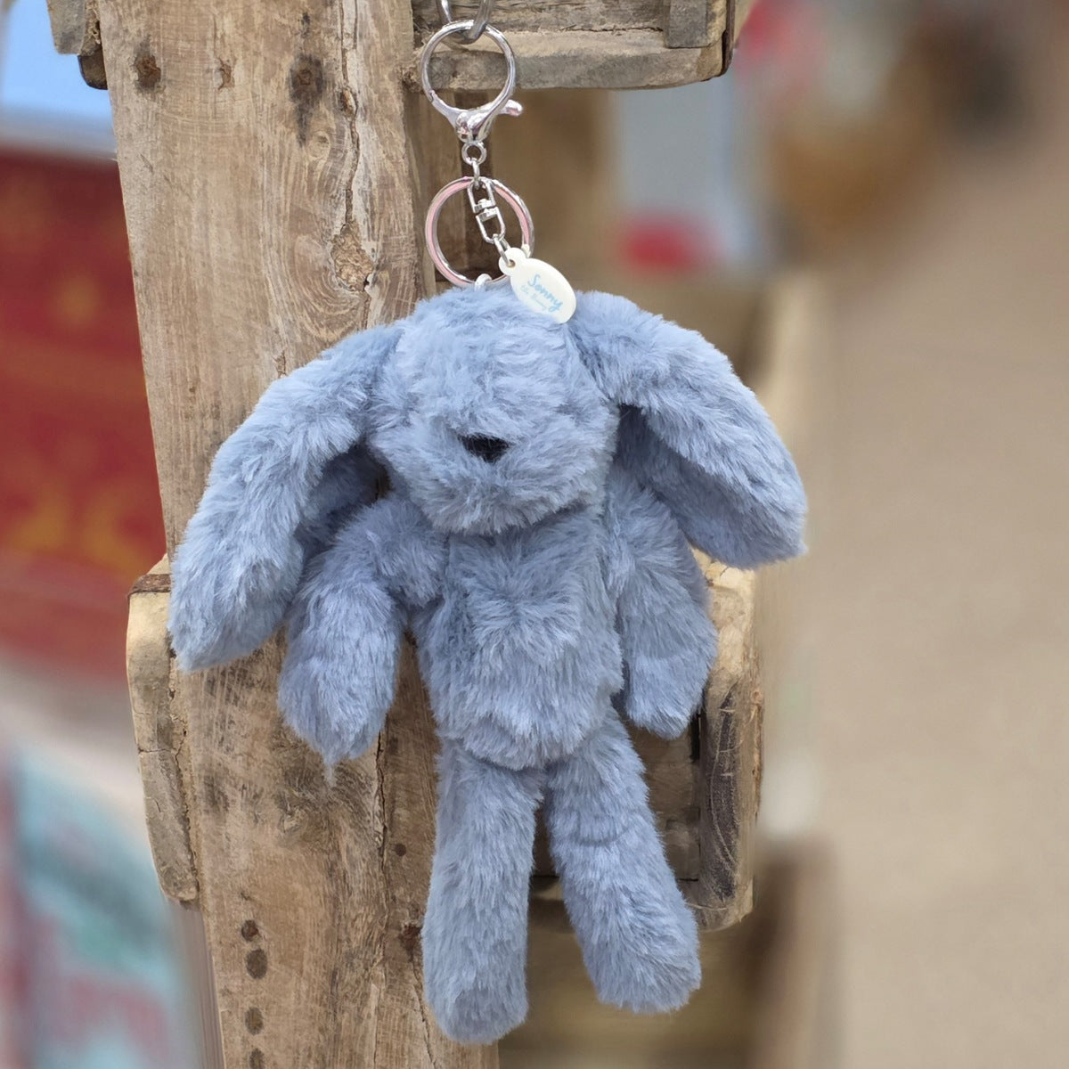 A blue plush bunny keychain hanging from a rustic wooden post. The keychain features a soft, fuzzy blue bunny with black eyes and nose, attached to a silver metal key ring and a small round tag.
