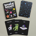 Space Snap card game set with the main box, the back of the card deck, and three sample cards showing space-themed illustrations on a light grey background.