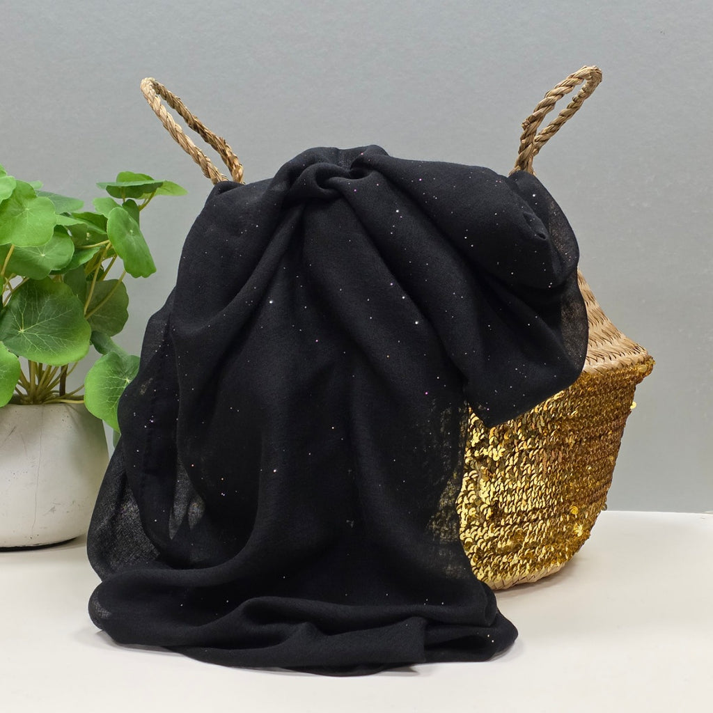 A black sheer fabric scarf with subtle silver glitter flecks, draped over a woven straw basket with natural fiber handles, with a potted green plant in the background.