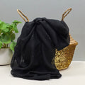 A black sheer fabric scarf with subtle silver glitter flecks, draped over a woven straw basket with natural fiber handles, with a potted green plant in the background.