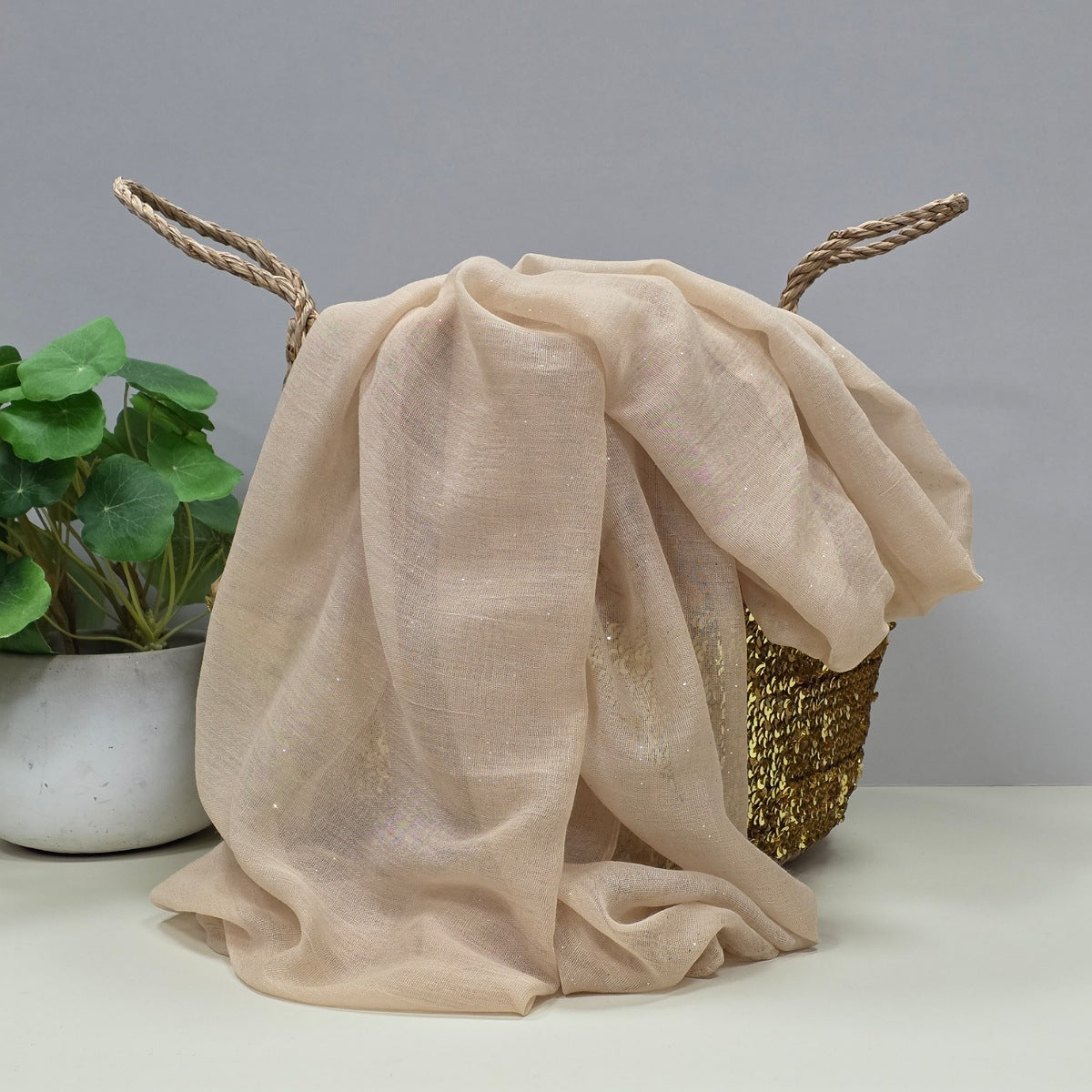 Beige sparkly sheer fabric scarf fabric draped over a basket with a plant on a gray background