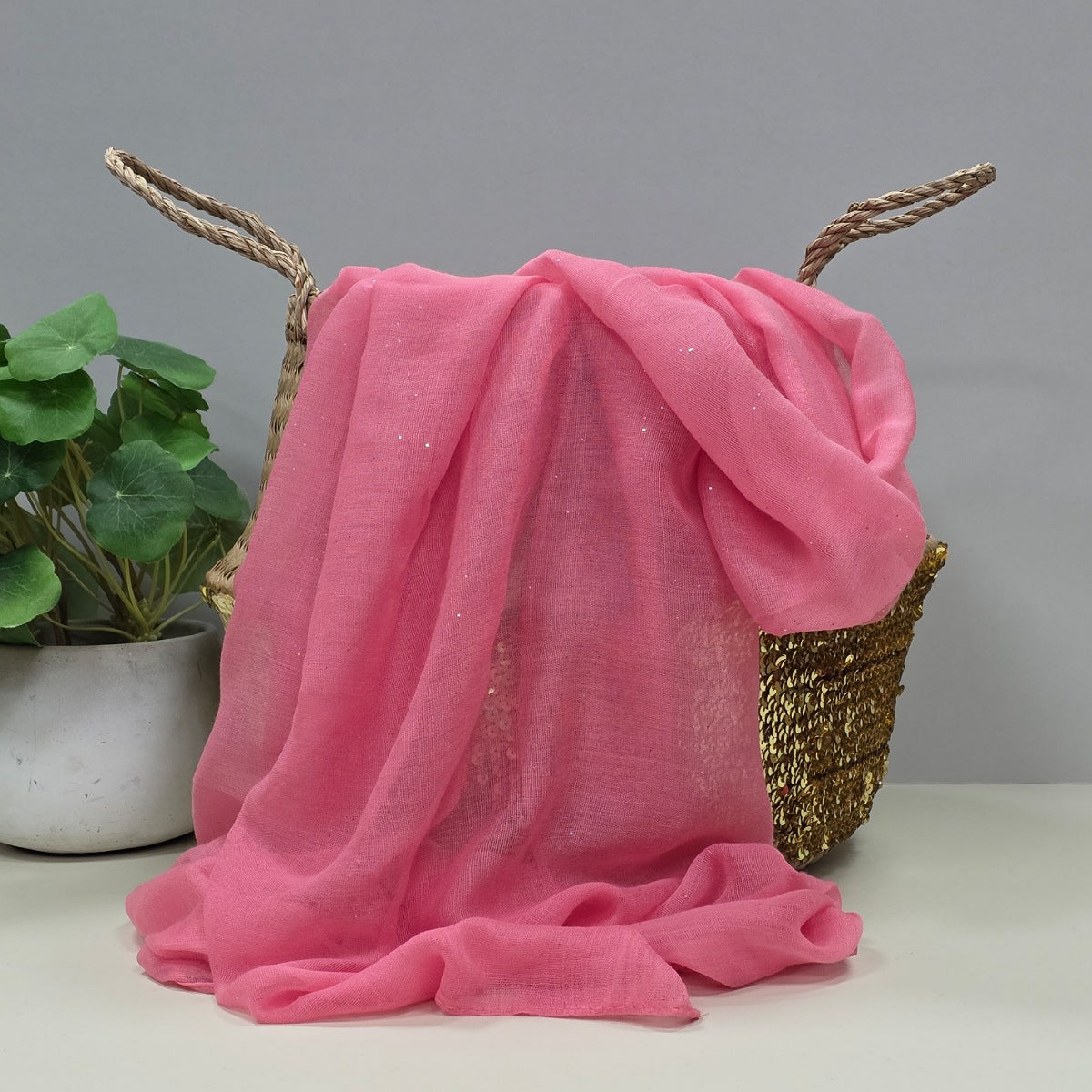 Dusky pink lightweight scarf with tiny sparkles draped over a decorative basket with a plant in the background
