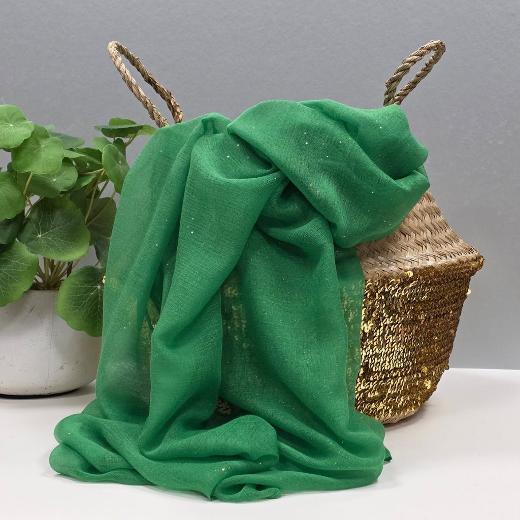 Grass green lightweight scarf with shiny sparkle detail draped over a woven basket with a plant on a grey background