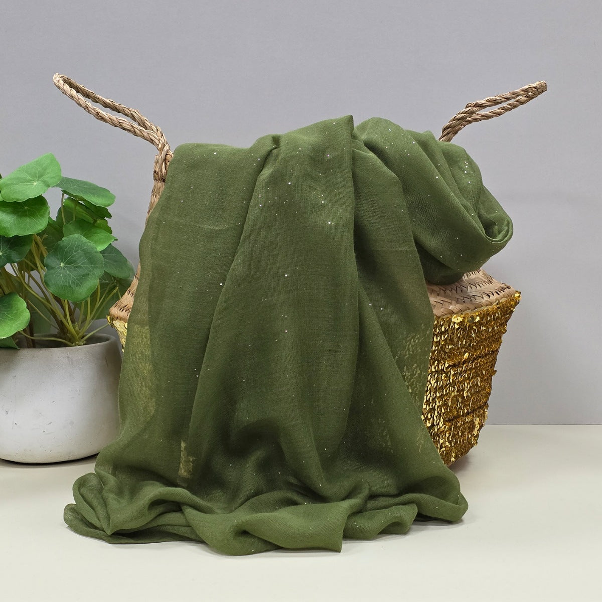 Khaki green lightweight scarf with gentle sparkle draped over a wicker basket with a plant in the background