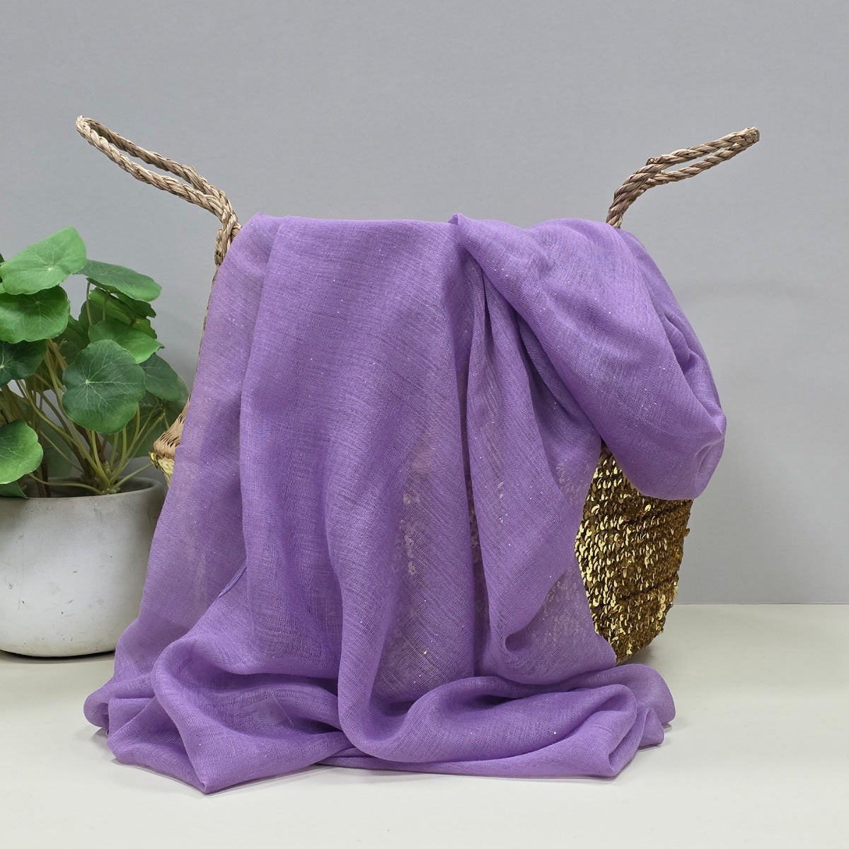 A lavender purple lightweight fabric scarf with gentle sparkles draped over a woven basket with rope handles, set on a white surface next to a potted green plant against a light grey background.
