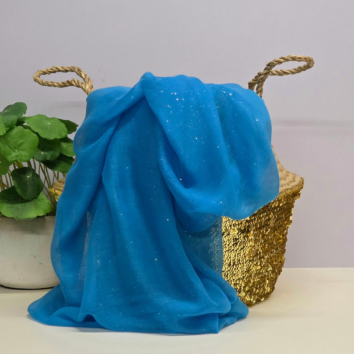 A bright blue sheer fabric scarf with tiny shiny speckles draped over a woven straw basket with rope handles, next to a potted green plant on a white surface against a light grey background.