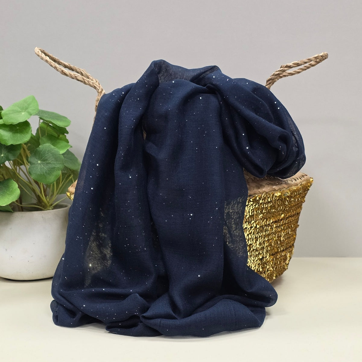 Navy blue fabric lightweight scarf with sparkle decoration draped over a basket on a light surface with a plant in the background.