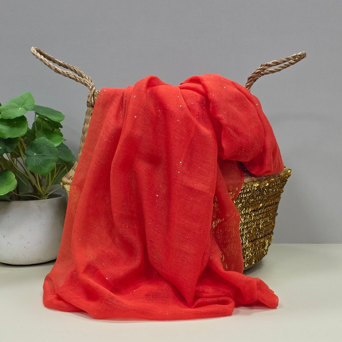 A bright red sheer fabric scarf with subtle glitter, draped over a woven gold basket with rope handles, next to a potted green plant on a light grey background.