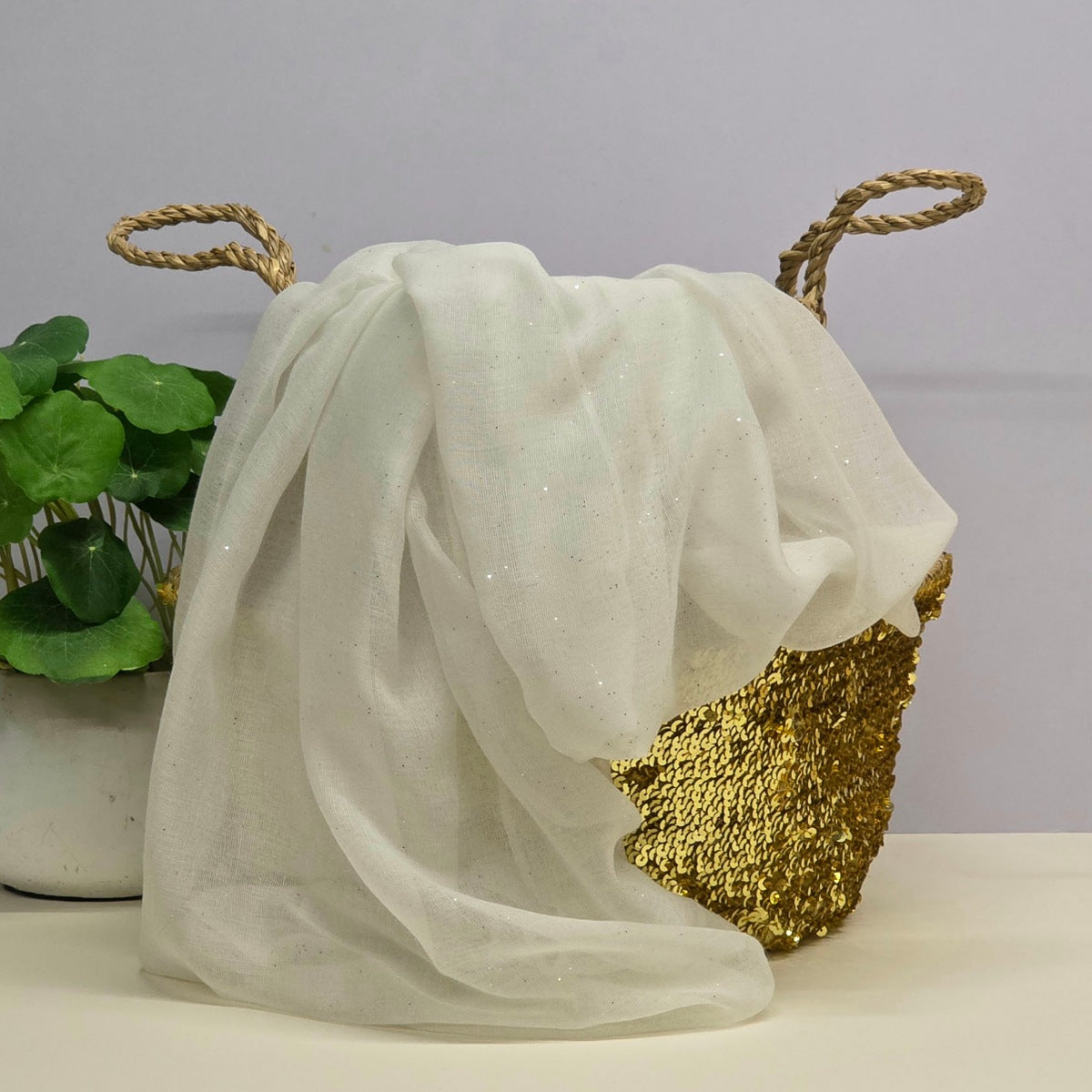 A white sheer tulle fabric with subtle glitter, draped over a gold sequined basket with rope handles, next to a potted green plant on a light surface against a pale gray background.