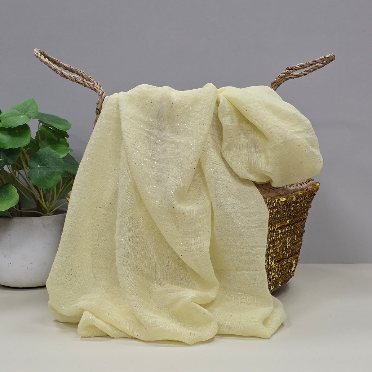 A pale yellow sheer fabric with subtle glittery flecks draped over a woven basket, with a potted green plant in the background against a neutral grey wall.