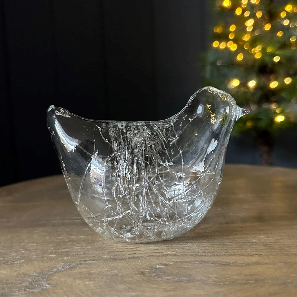 A clear glass bird-shaped ornament with a textured, spun glass pattern, resting on a wooden tabletop with a blurred Christmas tree in the background.