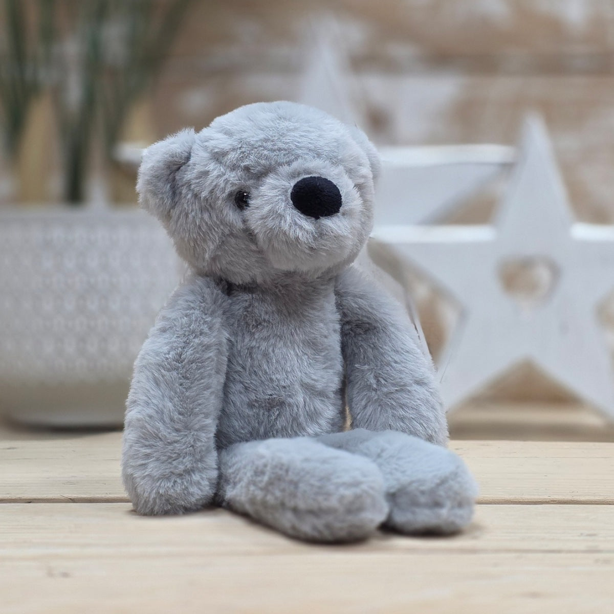 A light grey soft fuzzy plush teddy bear toy, with black eyes and nose, on a wooden surface with a blurred star-shaped object in the background
