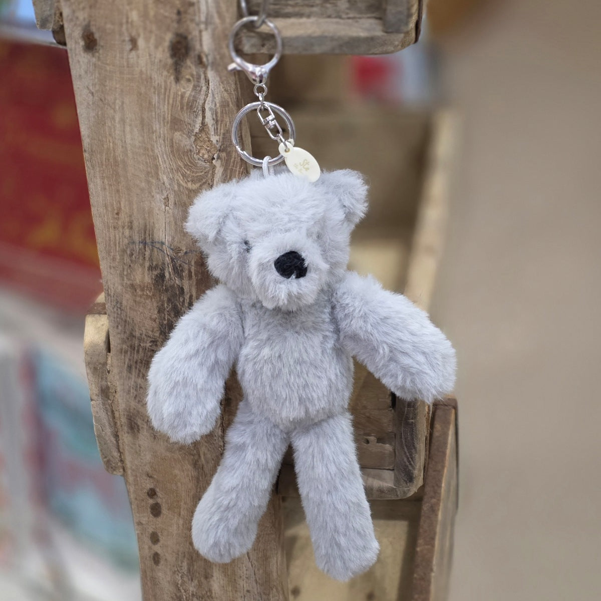 A light grey plush teddy bear keychain hanging from a rustic wooden post. The keychain features a soft, fuzzy grey bear with black eyes and nose, attached to a silver metal key ring and a small round tag.
