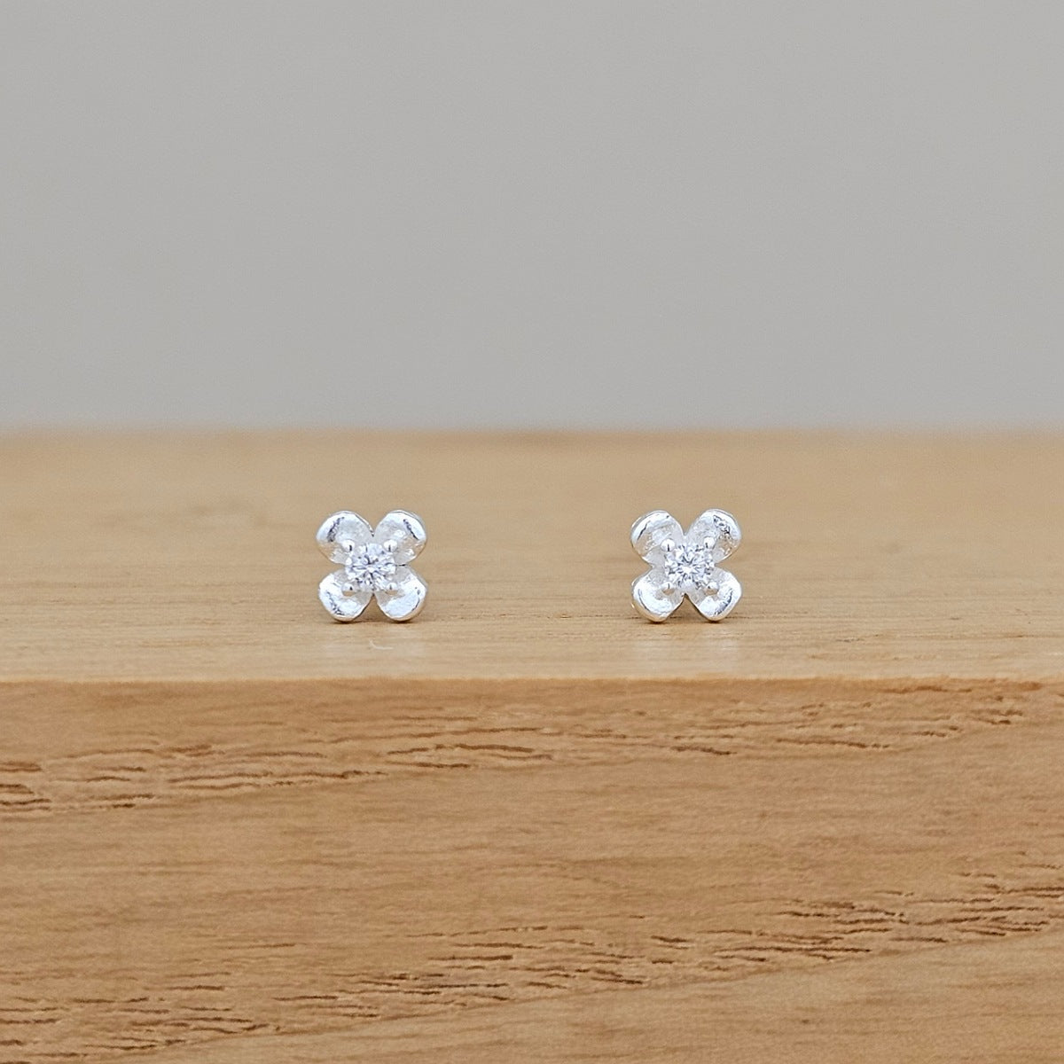 A pair of silver four-petal flower stud earrings with clear stones, resting on a light brown wooden surface against a neutral gray background.