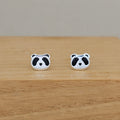 A pair of silver stud earrings shaped like panda faces, featuring black enamel details, displayed on a light wooden surface against a neutral beige background.