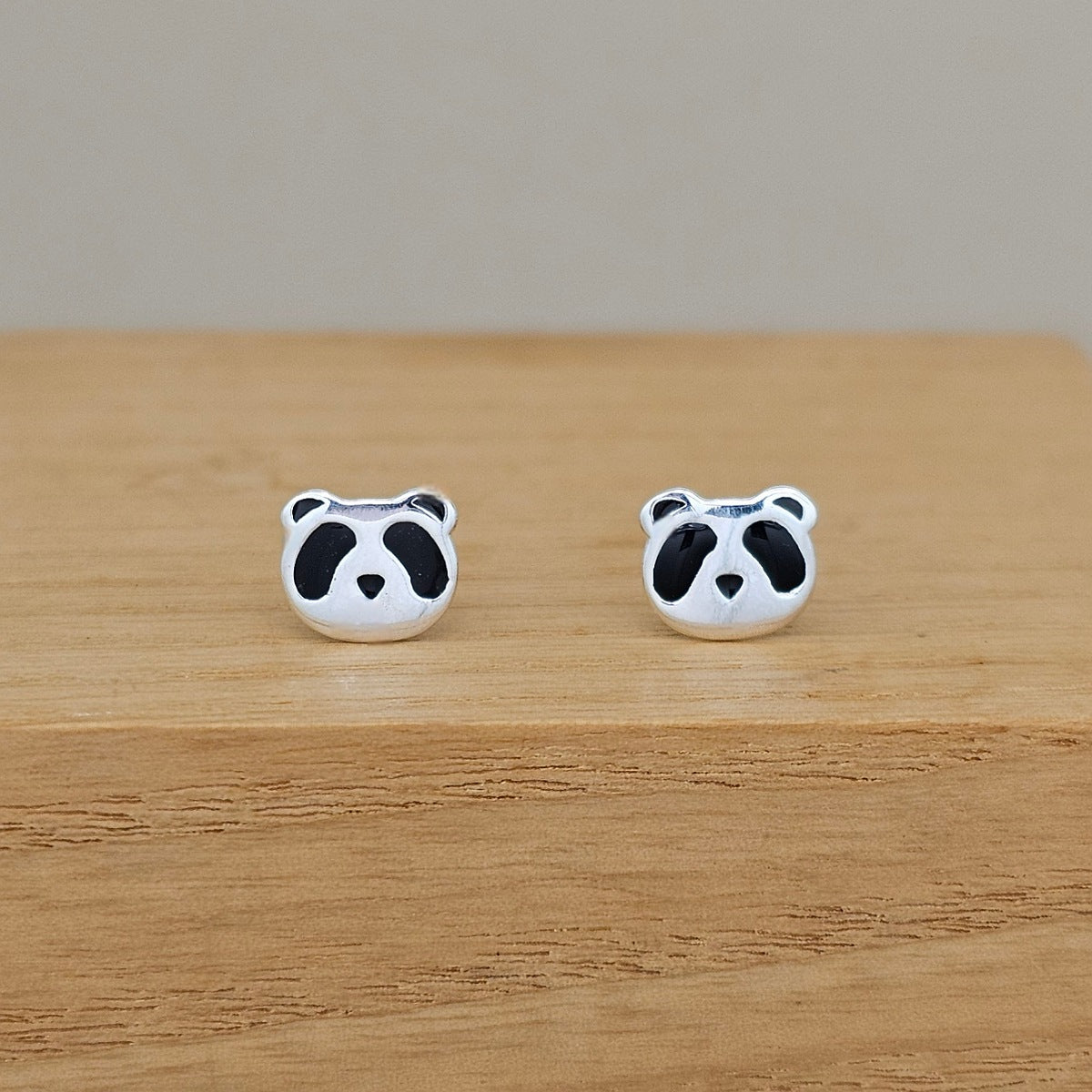 A pair of silver stud earrings shaped like panda faces, featuring black enamel details, displayed on a light wooden surface against a neutral beige background.