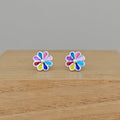 A pair of silver flower stud earrings with multicolored enamel petals, resting on a light wood surface against a neutral gray background.