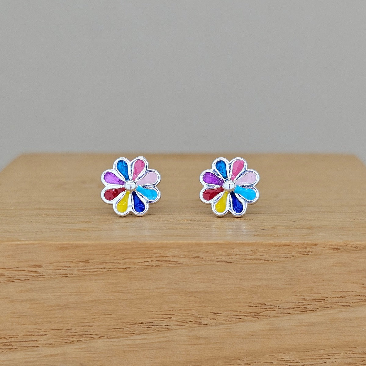 A pair of silver flower stud earrings with multicolored enamel petals, resting on a light wood surface against a neutral gray background.