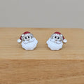 A pair of Santa Claus stud earrings with white enamel beard, silver-tone metal face and hat trim, and a red hat, displayed on a light wood surface.