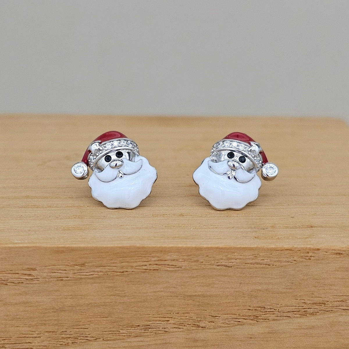 A pair of Santa Claus stud earrings with white enamel beard, silver-tone metal face and hat trim, and a red hat, displayed on a light wood surface.