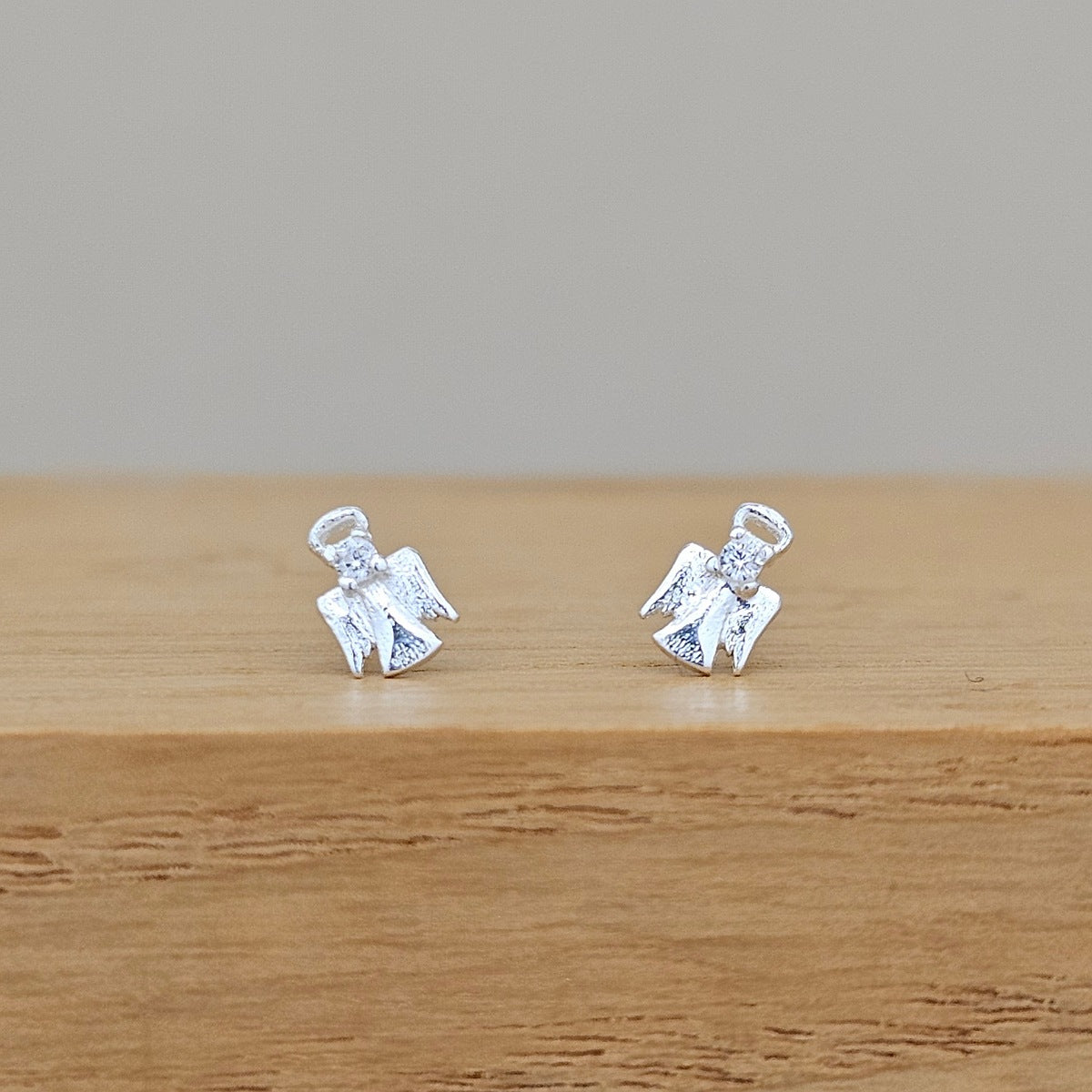 A pair of small silver angel stud earrings with textured wings and halo details, displayed on a wooden surface against a neutral gray background.