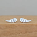 A pair of silver angel wing stud earrings resting on a light brown wooden surface against a neutral gray background.