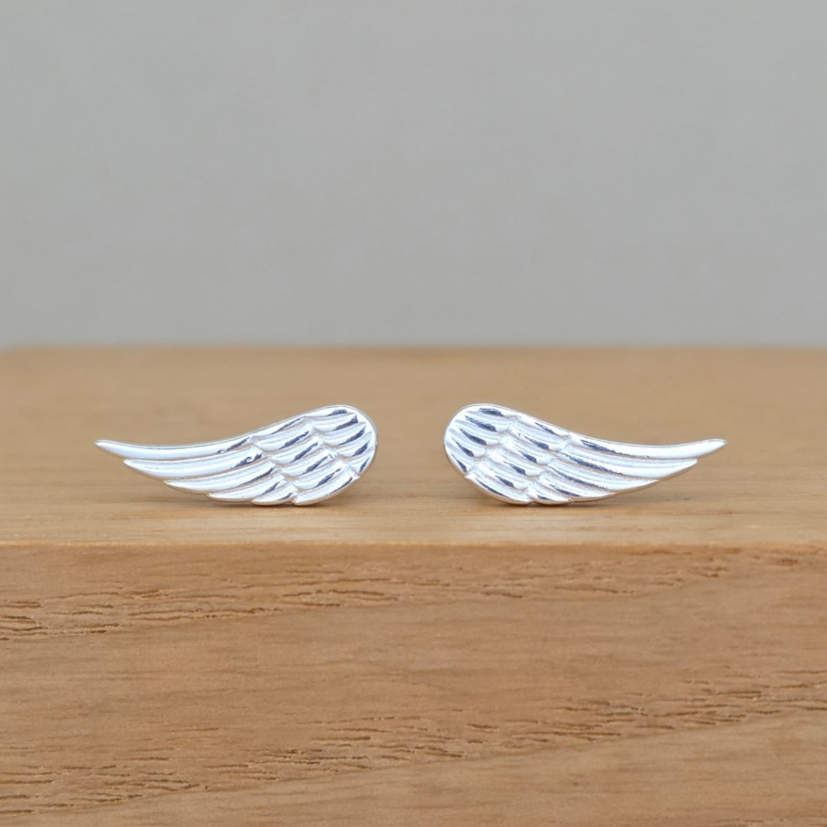 A pair of silver angel wing stud earrings resting on a light brown wooden surface against a neutral gray background.