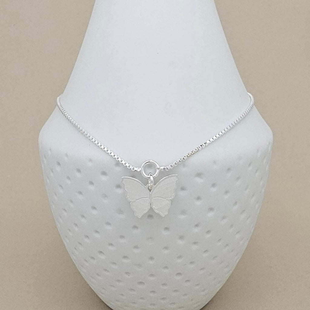 A delicate silver chain anklet with a small butterfly pendant, displayed on a textured white jewelry bust against a neutral beige background.