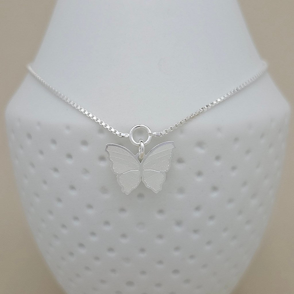 A delicate silver chain anklet with a small butterfly pendant, displayed on a white textured bust against a light beige background.