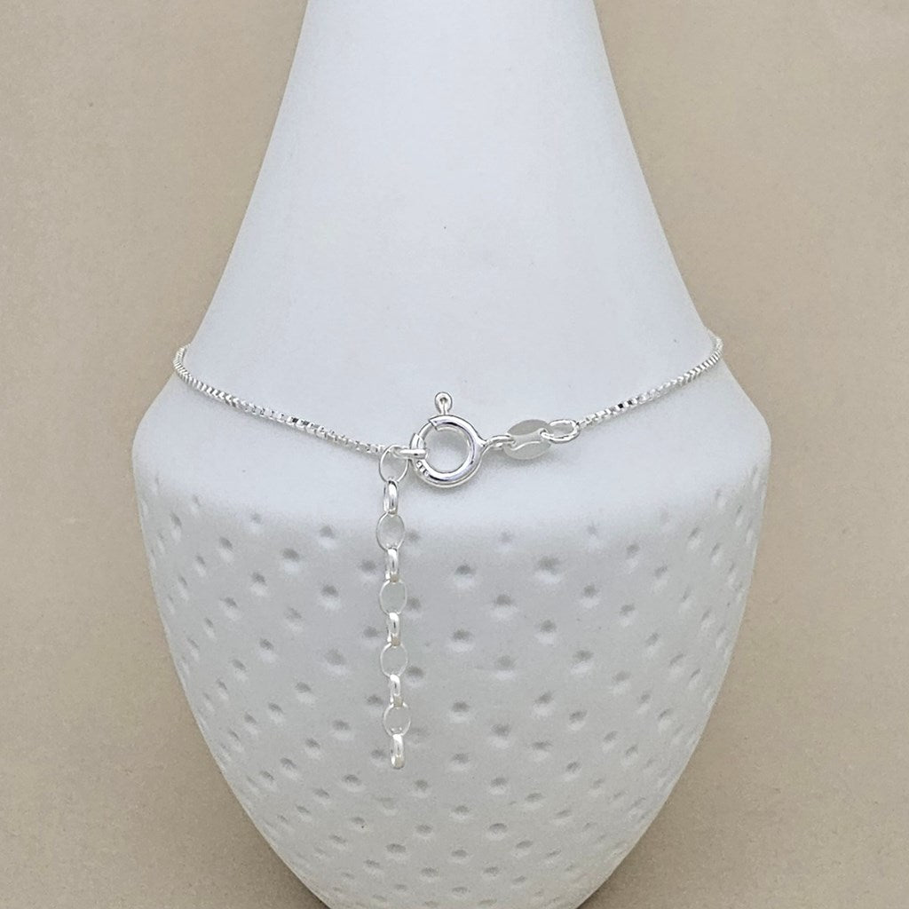 A delicate silver chain anklet with a circular ring clasp and extender chain, displayed on a white textured jewelry bust against a beige background.
