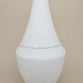 A delicate silver chain anklet displayed on a white ceramic cone-shaped jewelry stand against a solid beige background.