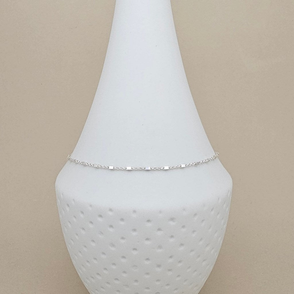 A delicate silver chain anklet displayed on a white ceramic cone-shaped jewelry stand against a solid beige background.