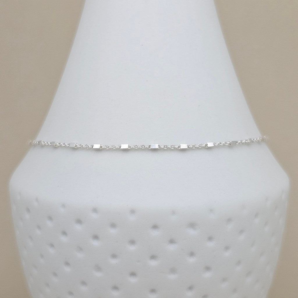 A delicate silver chain anklet with small rectangular links, displayed on a white textured cone jewelry stand against a light beige background.