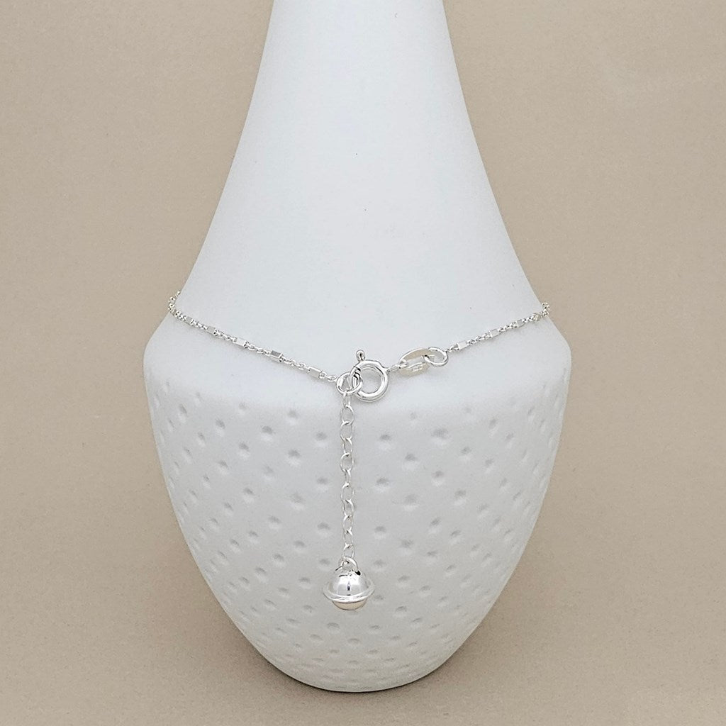 A delicate silver chain anklet with a small dangling ball charm, displayed on a white textured cone jewelry stand against a solid beige background.