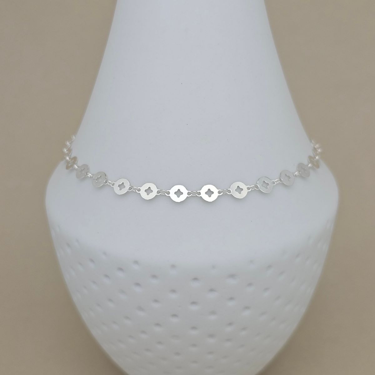 A delicate silver chain anklet featuring small, repeating circular medallion charms with a cross in the center, displayed on a white textured cone stand against a solid beige background.
