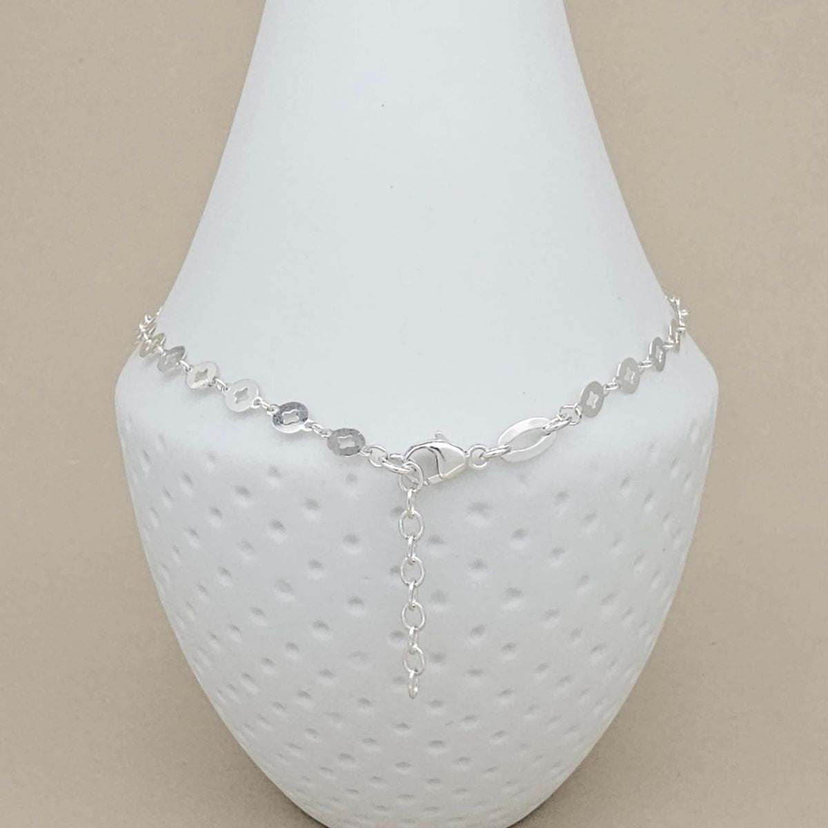 A silver chain anklet with oval links and a dangling charm, displayed on a white textured jewelry bust against a beige background.